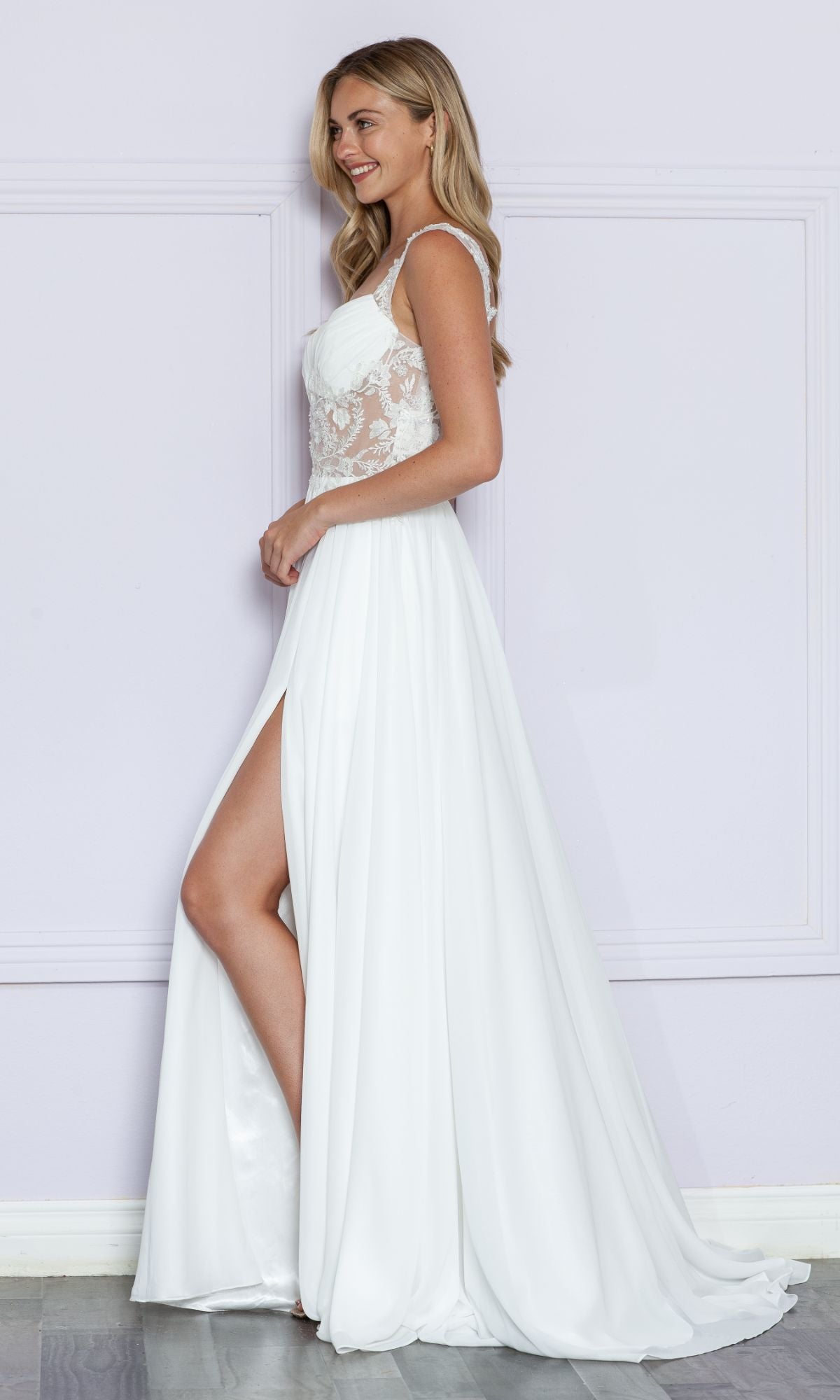 A woman models the Sheer-Lace Bodice Long White Formal Dress 9376, featuring a high slit, as she stands sideways against a light purple wall.